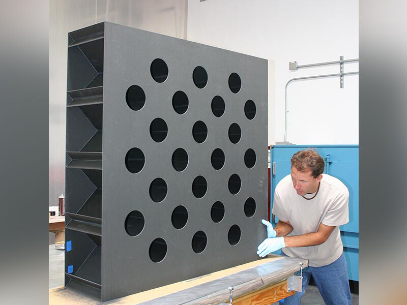 Exterior image of the near zero CTE antenna segment. Technician is adjusting the placement of the segment, which is on its side on a table in a manufacturing environment. The two exterior surface panels are sandwiched over the grid. The facing panel has circular holes in a grid pattern across its surface. The end of the grid section is visible along the edges between the two panels.